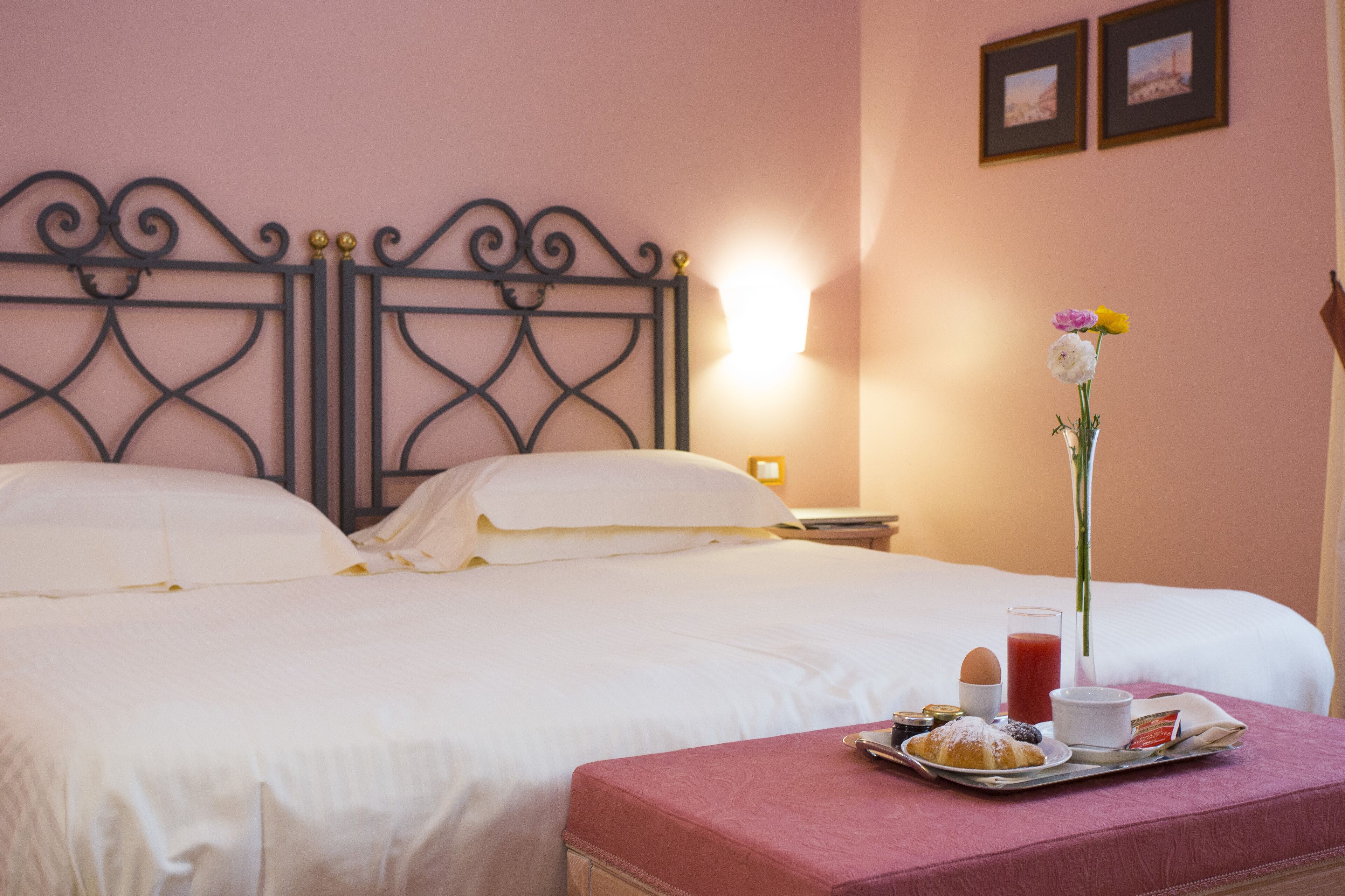 A neatly made bed with white linens, a tray with pastries, a glass of juice, and a vase with flowers on a pink ottoman.