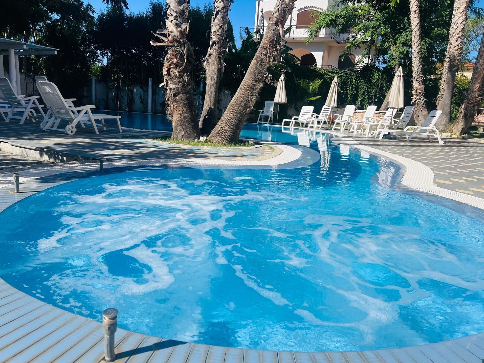 A swimming pool with lounge chairs and umbrellas, surrounded by palm trees and a building in the background.