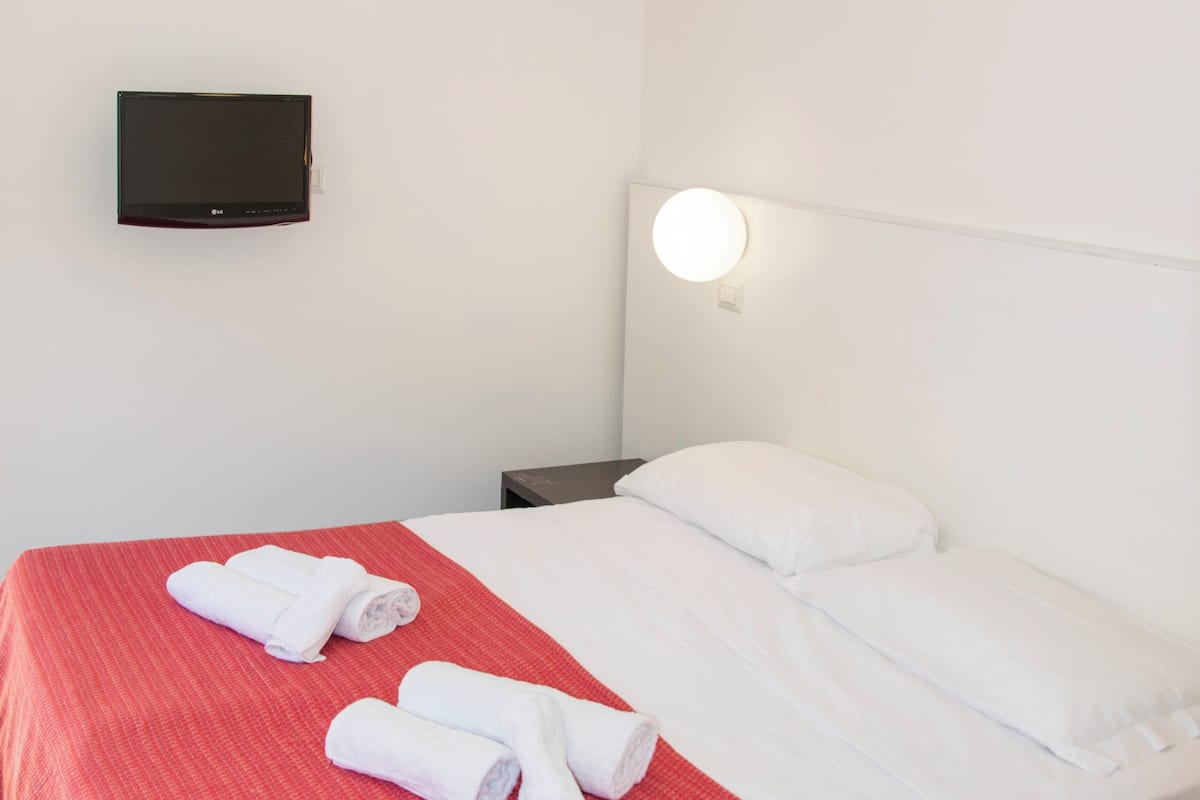 A hotel room with a bed, a flat-screen TV mounted on the wall, and a bedside table with a lamp.