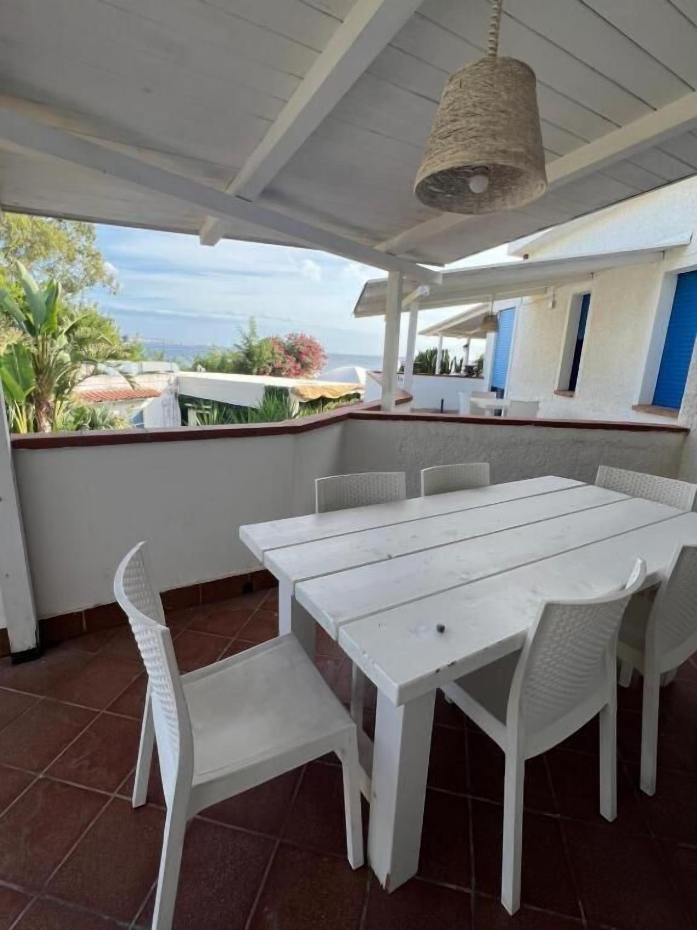 Un patio coperto con tavolo e sedie bianche, un lampadario e vista sul mare.