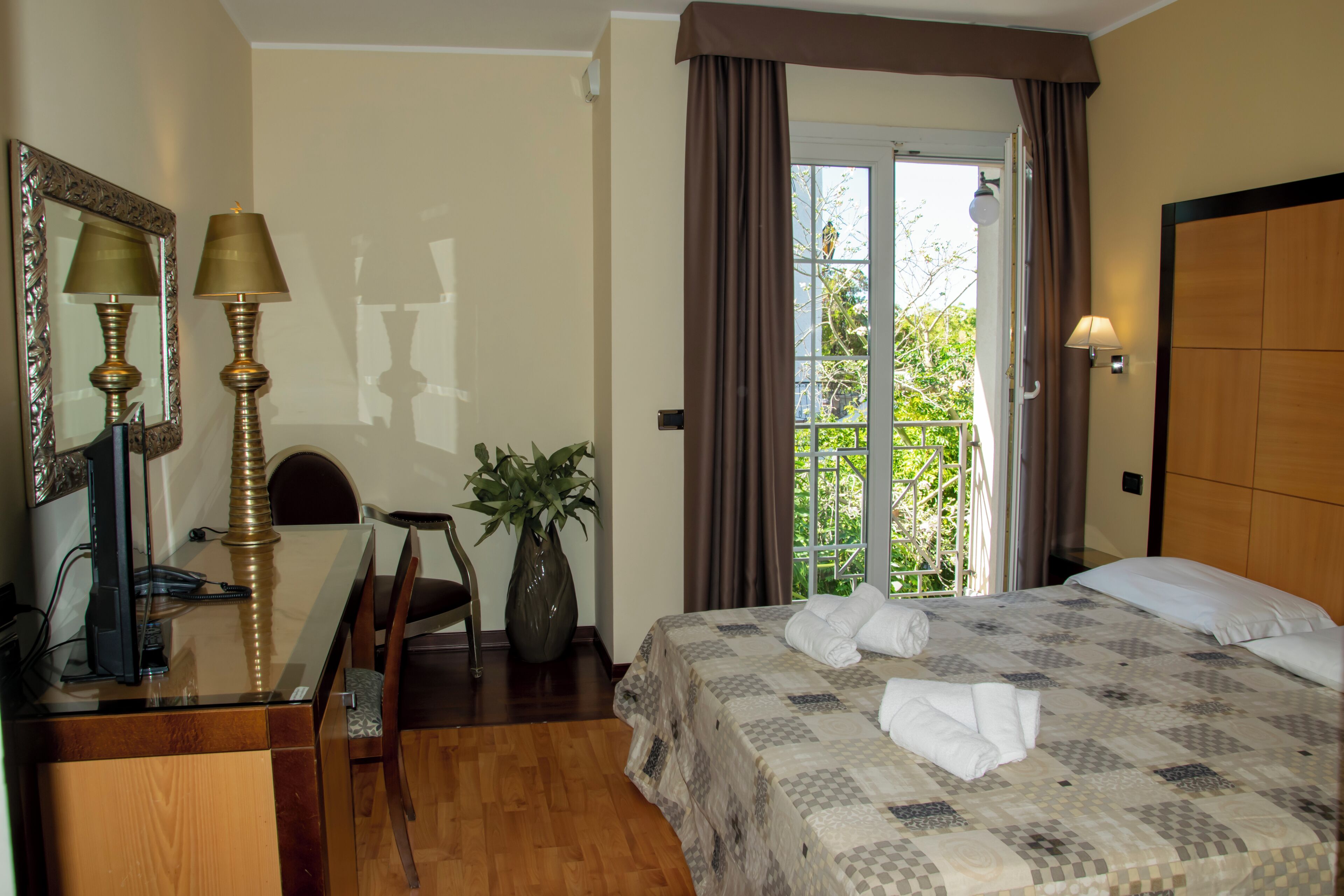 Une chambre d’hôtel avec un lit, un bureau doté d’une lampe, une chaise, une plante en pot et une vue sur l’extérieur.