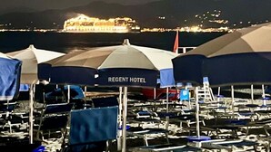 Vista desde la playa con varios sombrillas y sillas, un edificio iluminado a lo lejos, y una vista despejada del mar y las montañas.