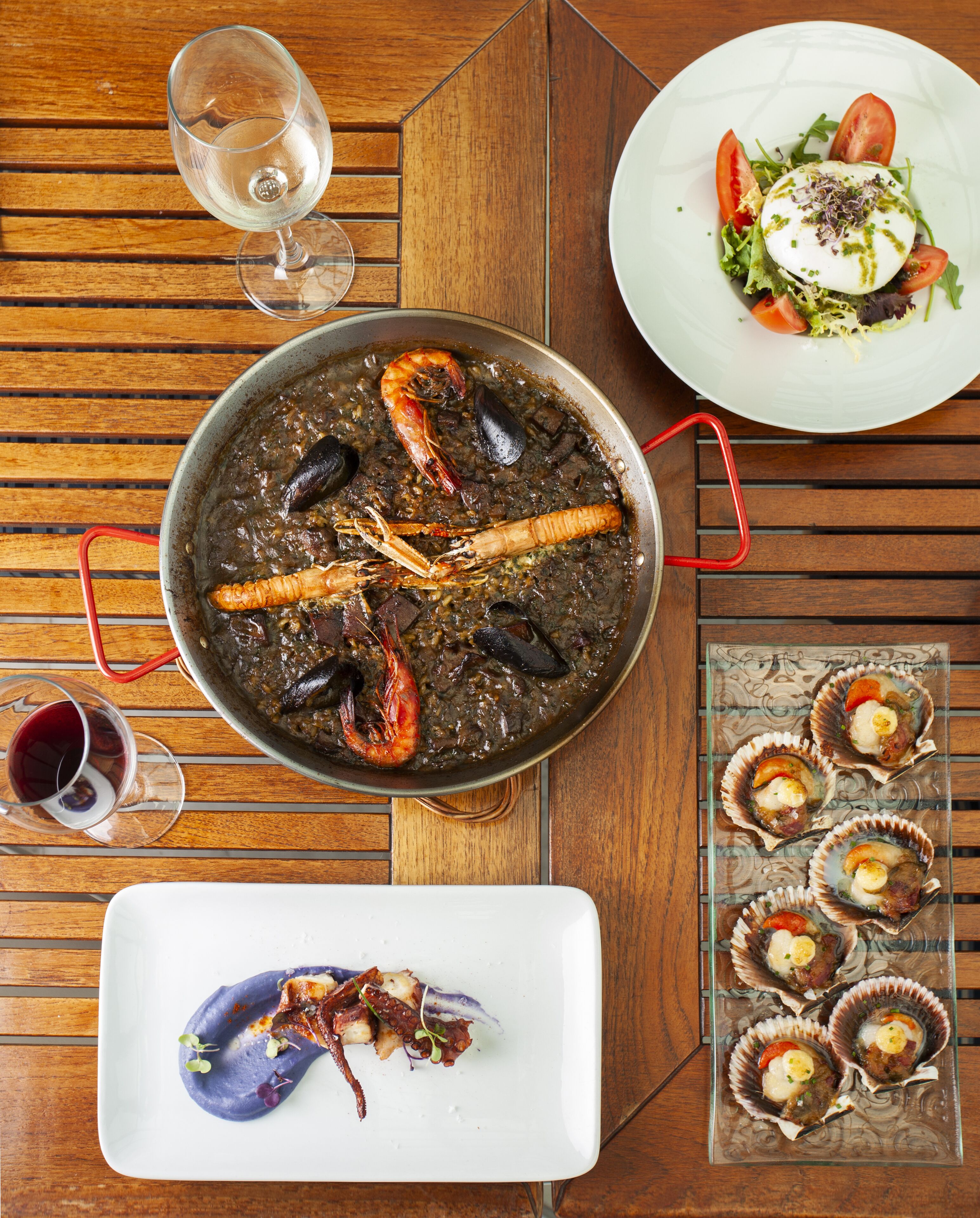 A dining table with a seafood dish, a glass of red wine, and a plate of scallops.