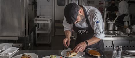 Un chef preparando comida en una cocina industrial con superficies de acero inoxidable, un teléfono y un carrito.