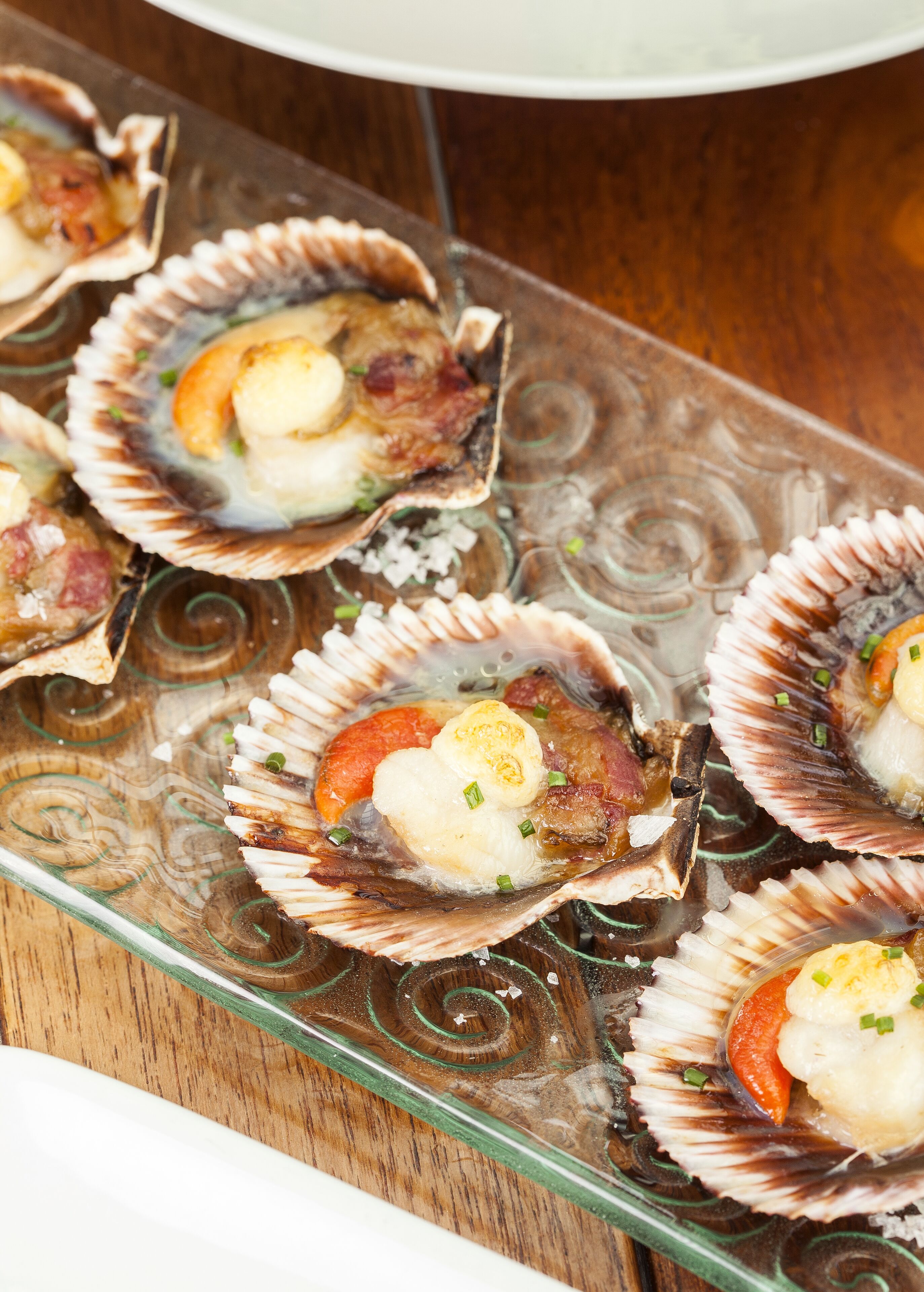 Un plateau en verre contenant des coquilles de Saint-Jacques, chacune garnie d’un morceau de viande, d’une tranche de tomate et d’une décoration.