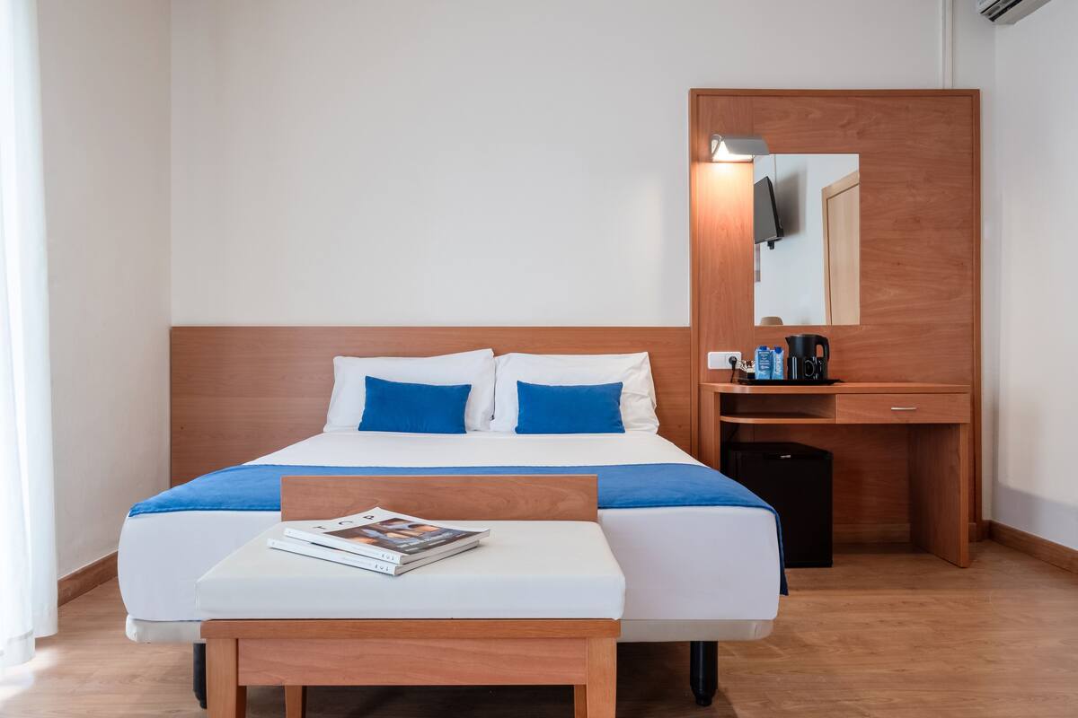 Ein Hotelzimmer mit einem Bett, einem Nachttisch mit einer Zeitschrift, einem Schreibtisch mit einer Kaffeemaschine und einem Spiegel.