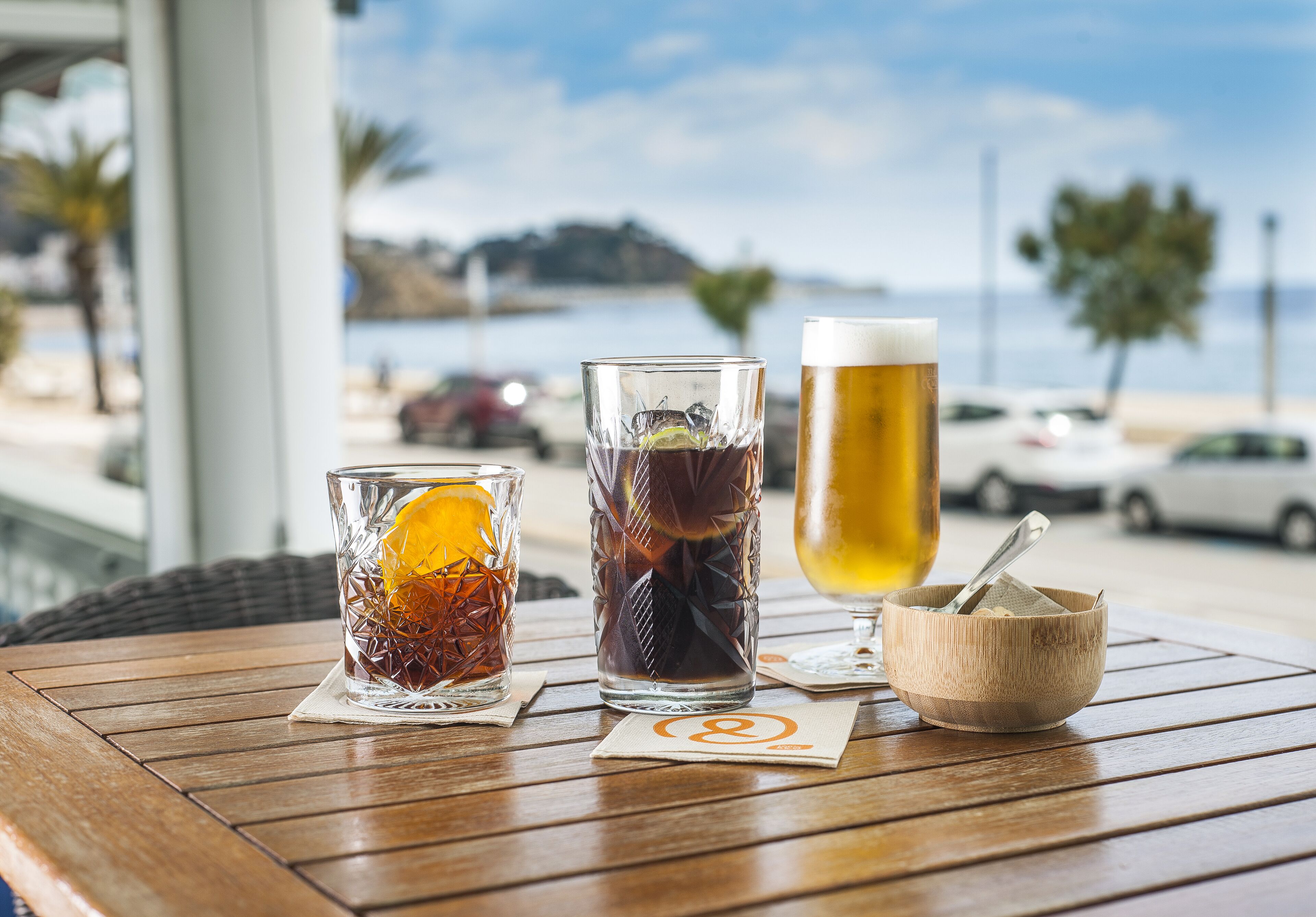 Ein Holztisch mit drei Gläsern und einer Schüssel, vor einer schönen Aussicht auf Strand und Autos.