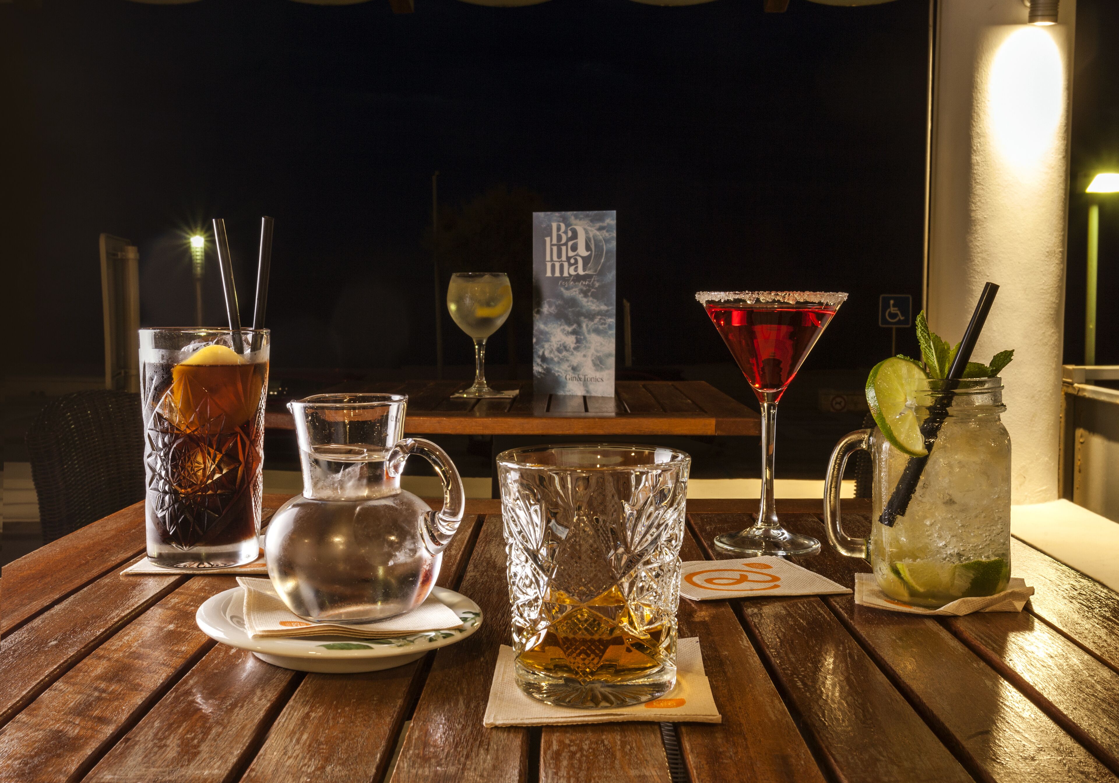 Een tafel met diverse drankjes, waaronder een hoog glas met een citroenpartje, een karaf, een glas met een limoen en een cocktailglas.