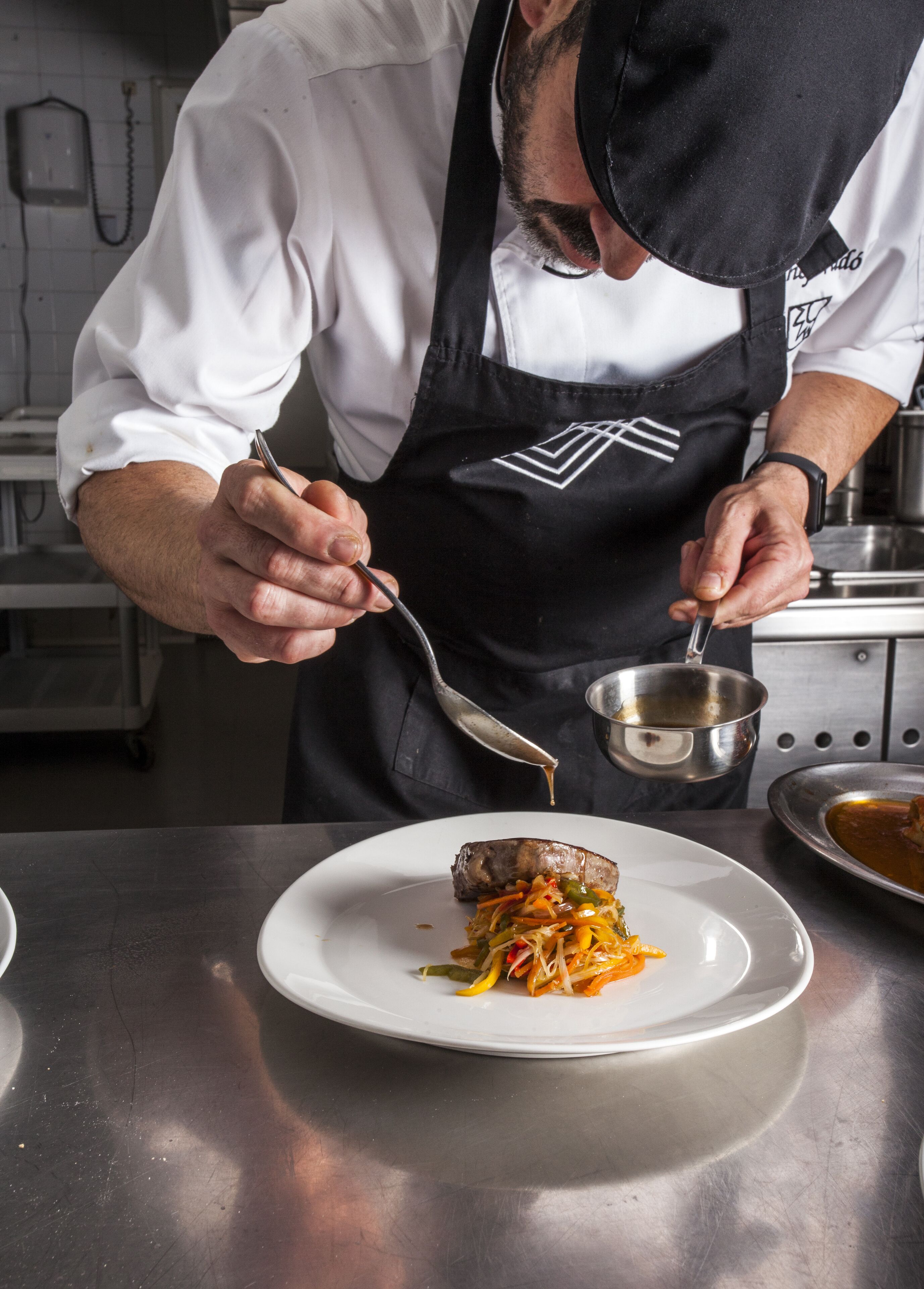 Un chef preparando un plato con una cuchara y un cuenco metálico pequeño.