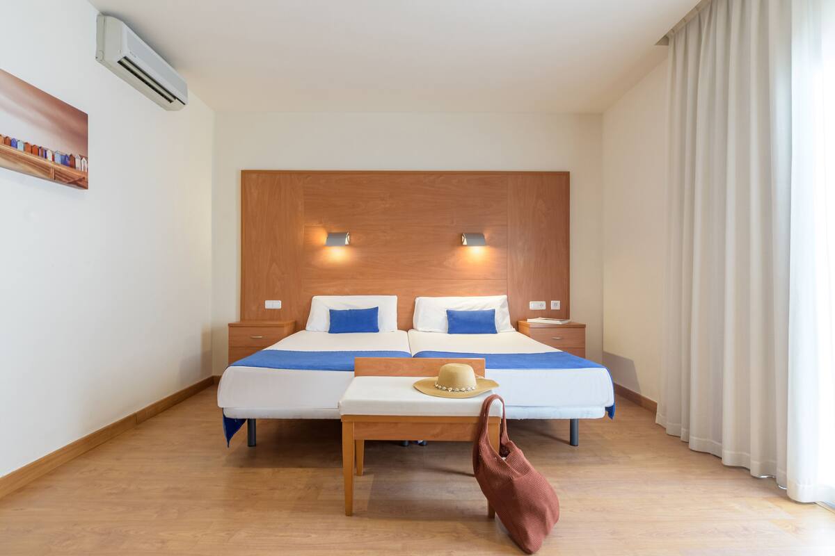 Ein Hotelzimmer mit Doppelbett, Nachttischen, einem Holzkopfende, einer Wandklimaanlage und einem Fenster mit Vorhängen.