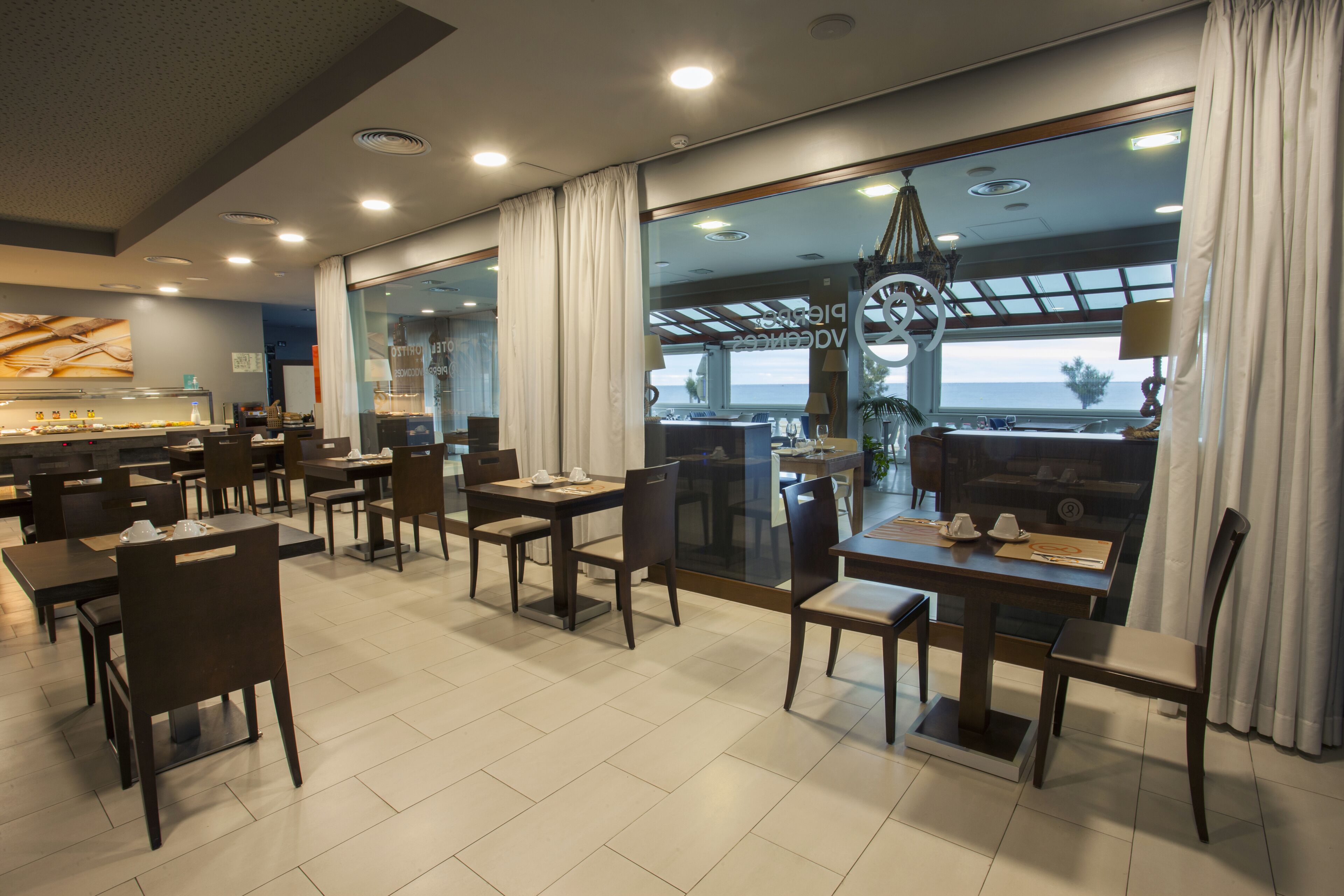 A modern dining area with tables and chairs, large windows with curtains, and a view of the sea.