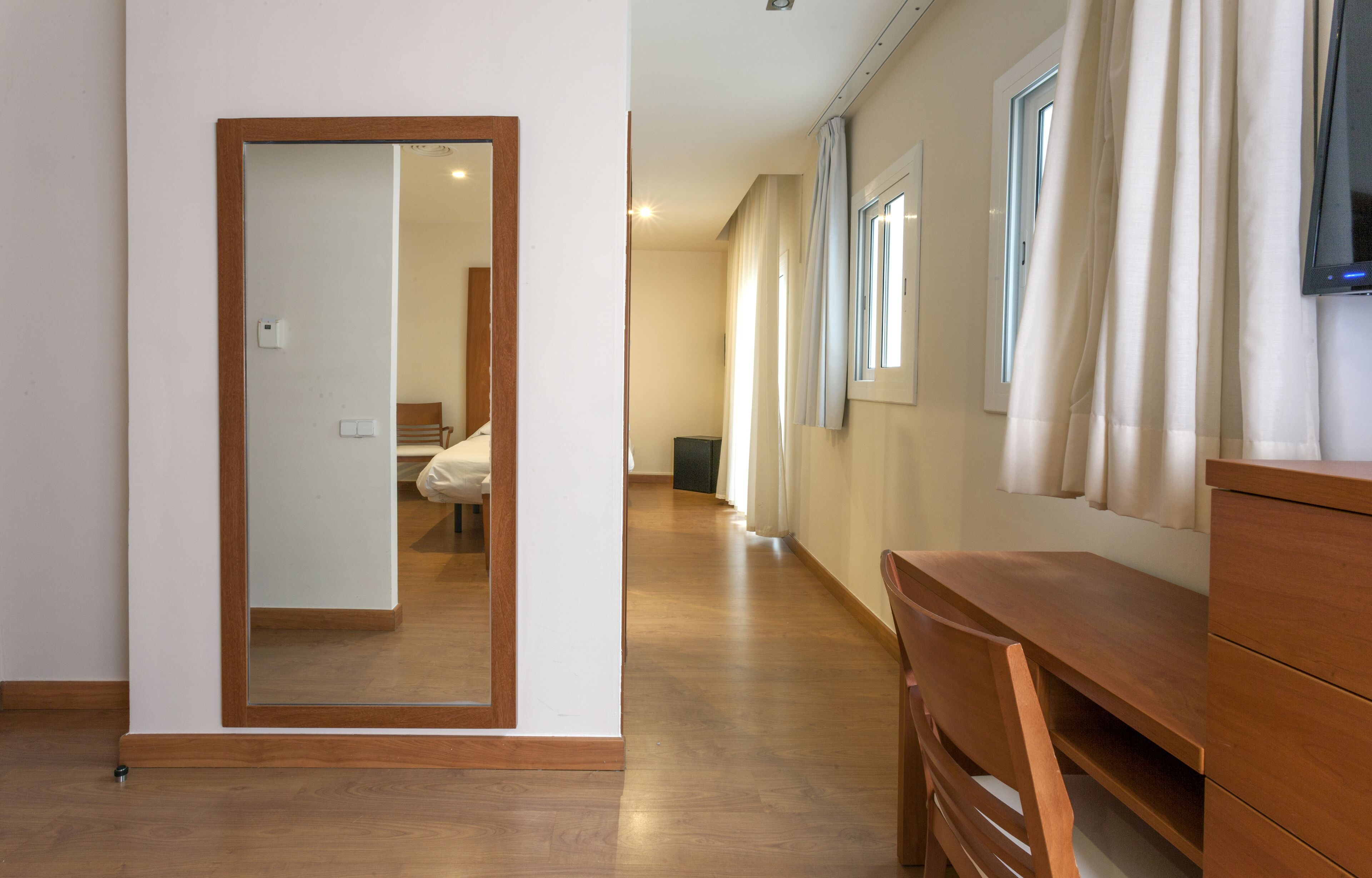 A hotel room with a wooden floor, a desk, a chair, a TV, and a window with curtains.