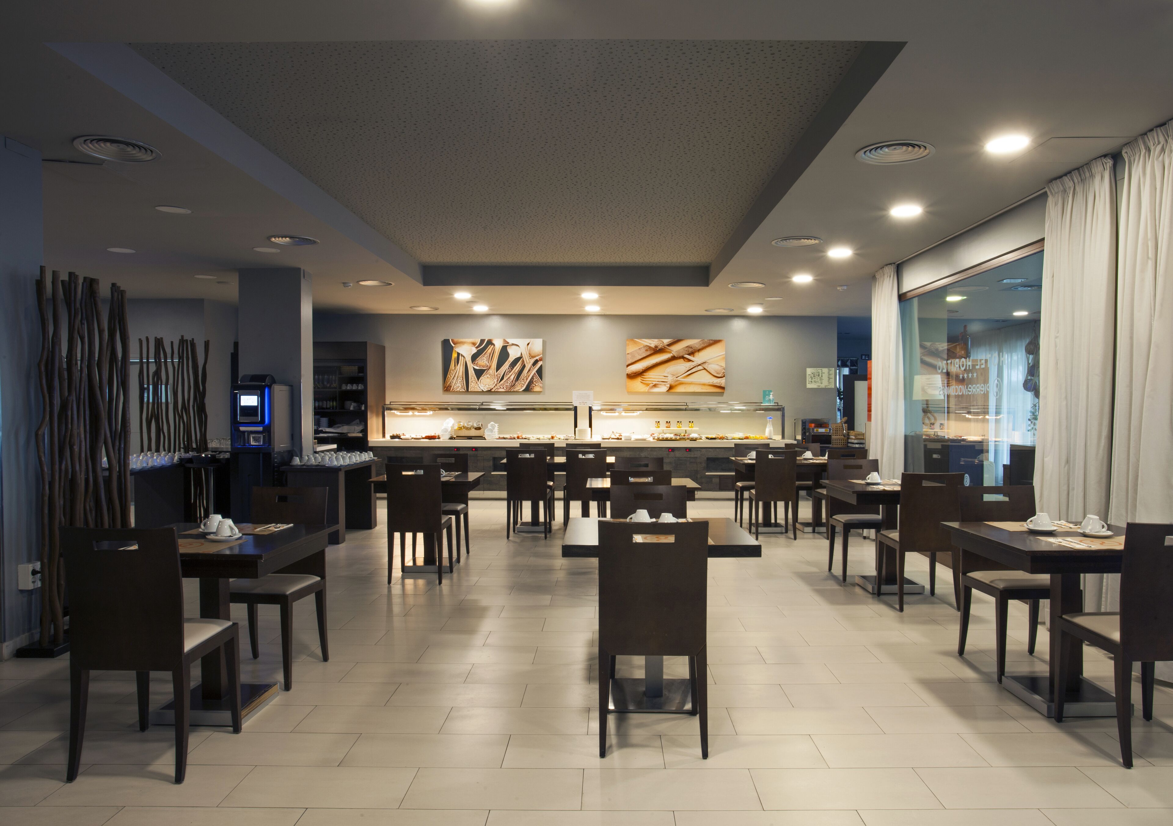 Un restaurant moderne doté d’un long comptoir, de nombreuses tables et des chaises. On y trouve des luminaires au plafond et une grande fenêtre agrémentée de rideaux.
