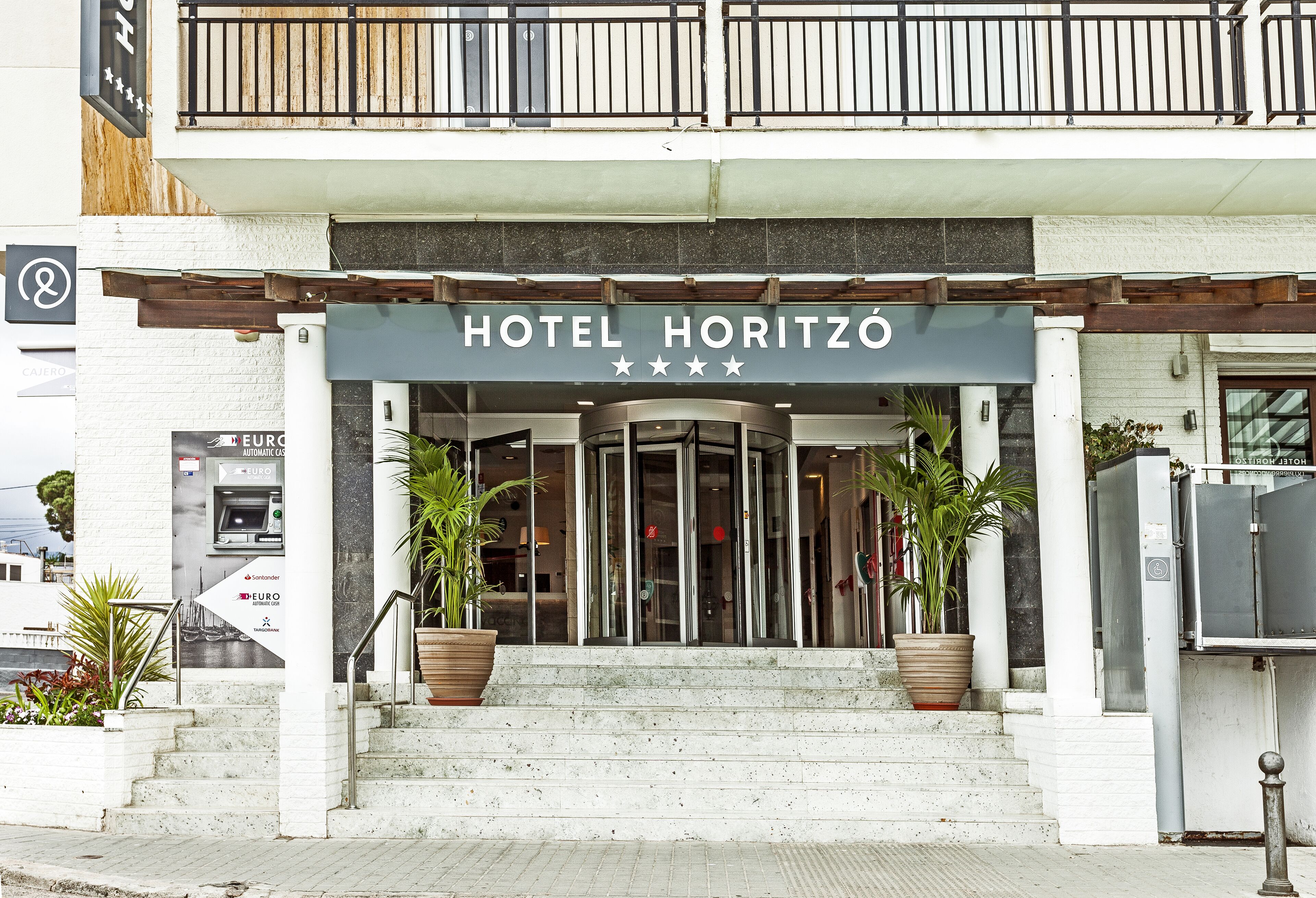 L'entrada d'HOTEL HORITZÓ amb un disseny modern, amb una porta de vidre, unes escales que condueixen a l'entrada i plantes en maceta a banda i banda.