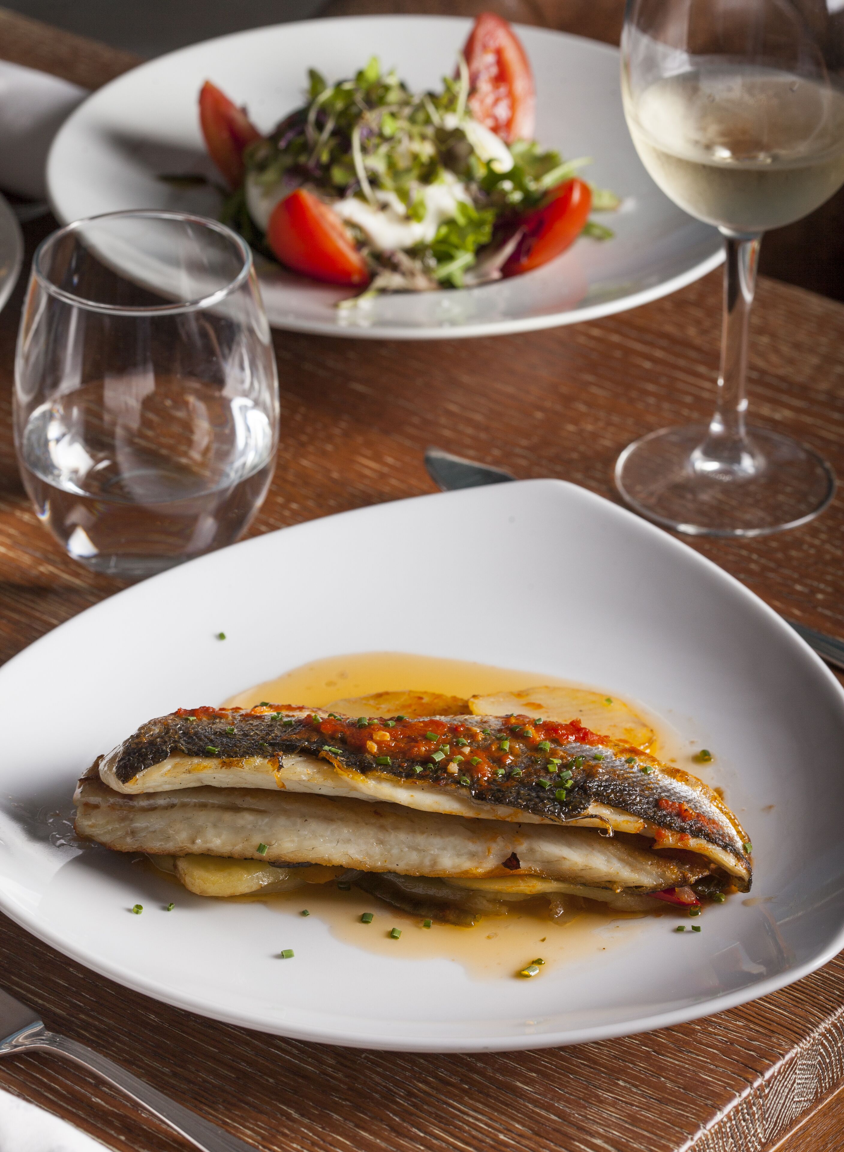 A plate of fish with a sauce and garnish, a glass of white wine, and a salad on a wooden table.