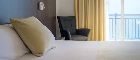 Habitación de hotel con cama, una silla, mesita de noche y vistas al mar a través de amplias ventanas.