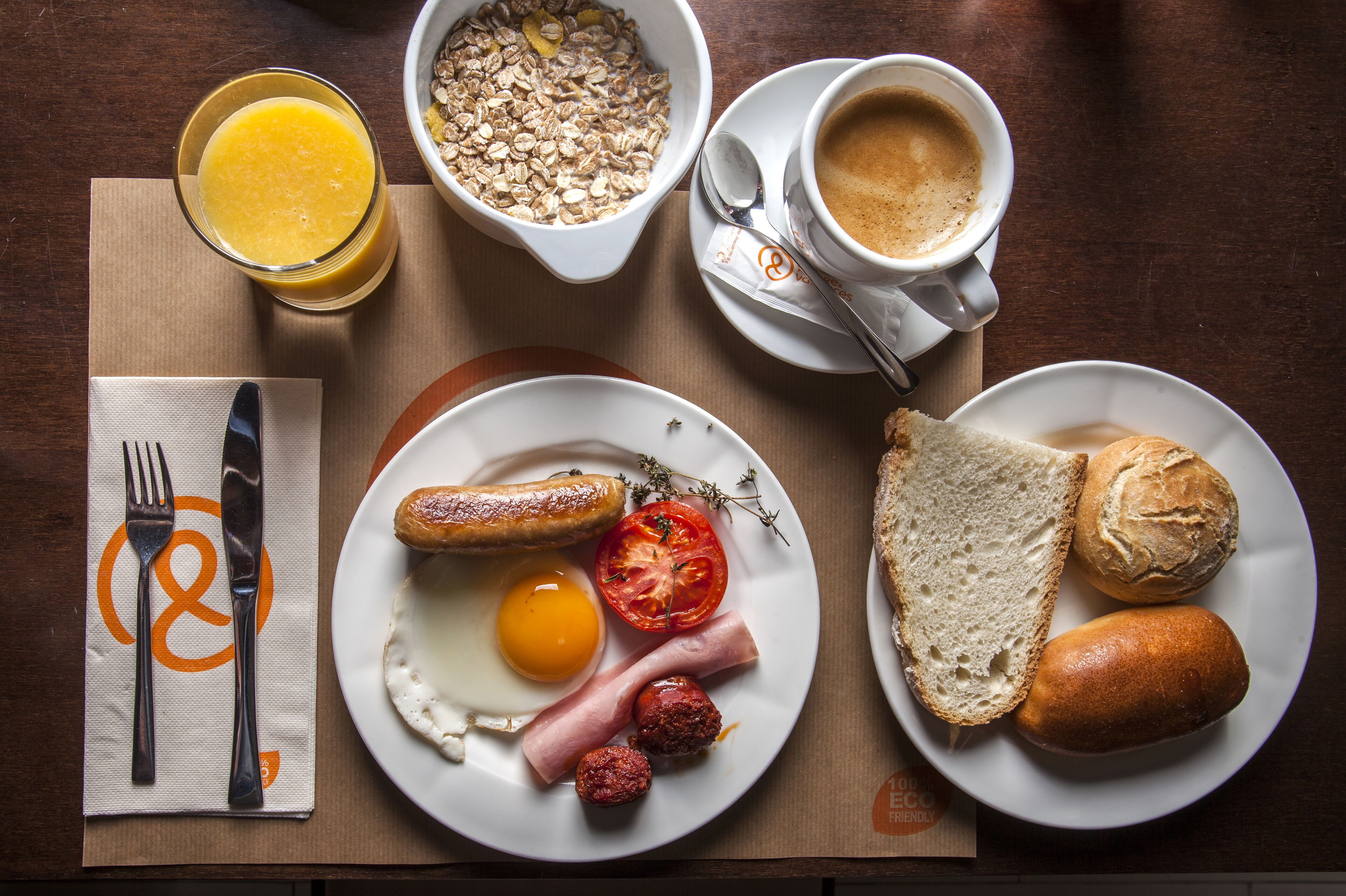 Ein Frühstückstisch mit Tellern voller Speisen, einem Orangensaftglas, einer Kaffeetasse und einer Serviette mit Gabel und Messer.