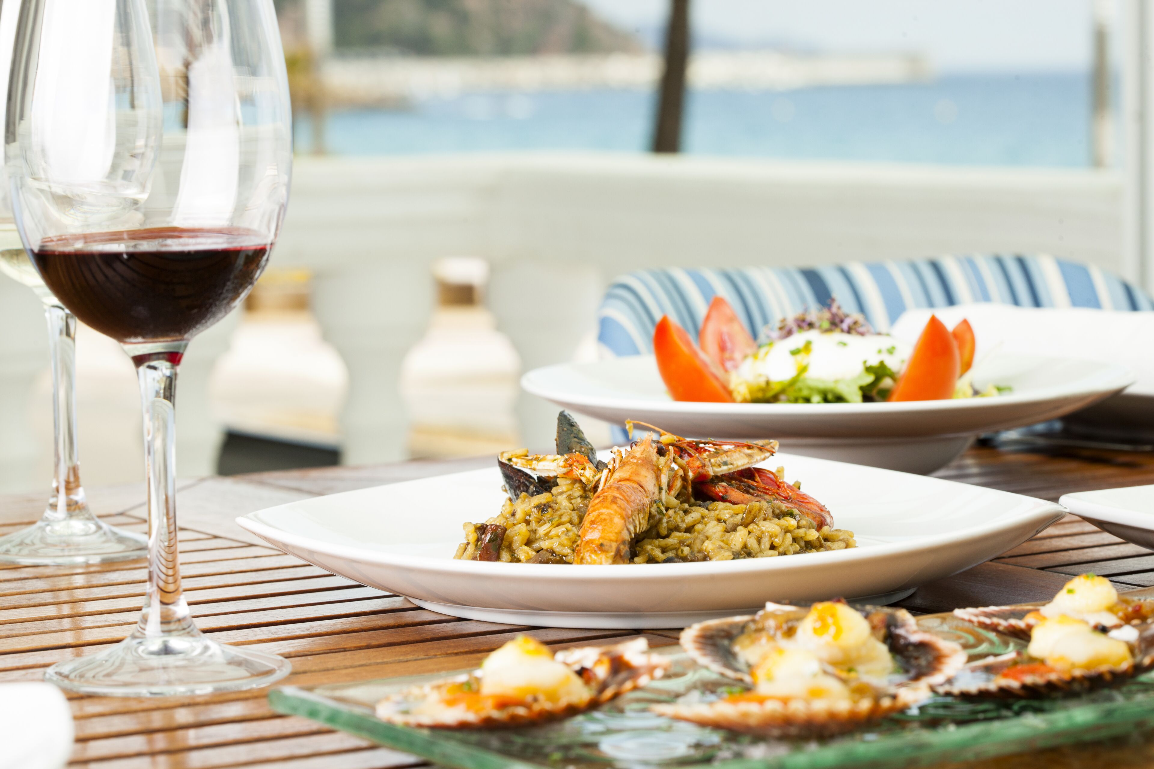Une table à manger dressée avec des plats de fruits de mer, un verre de vin rouge et une vue sur la mer.