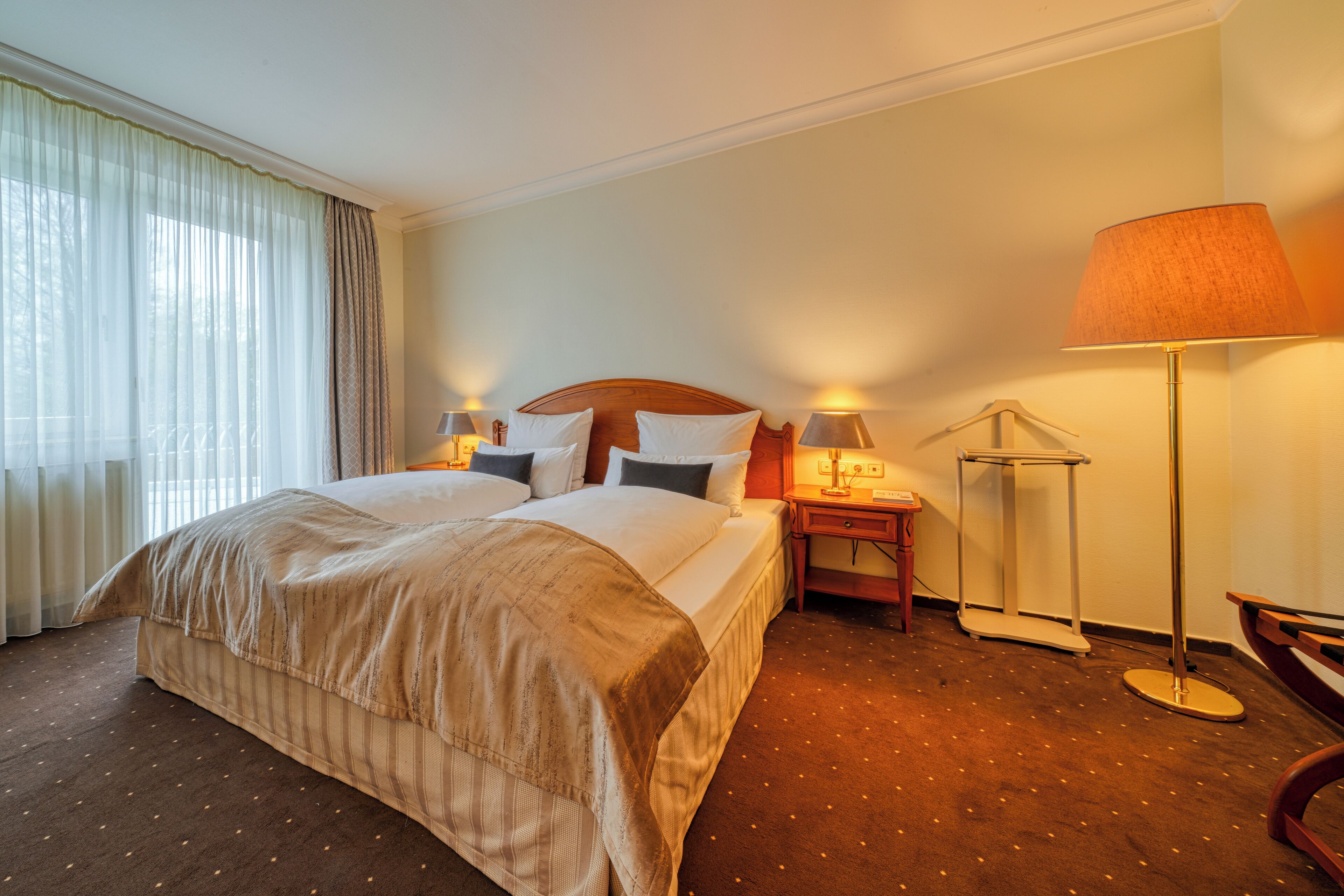Een hotelkamer met een groot bed, twee nachtkastjes met lampen, een vloerlamp en een bankje.