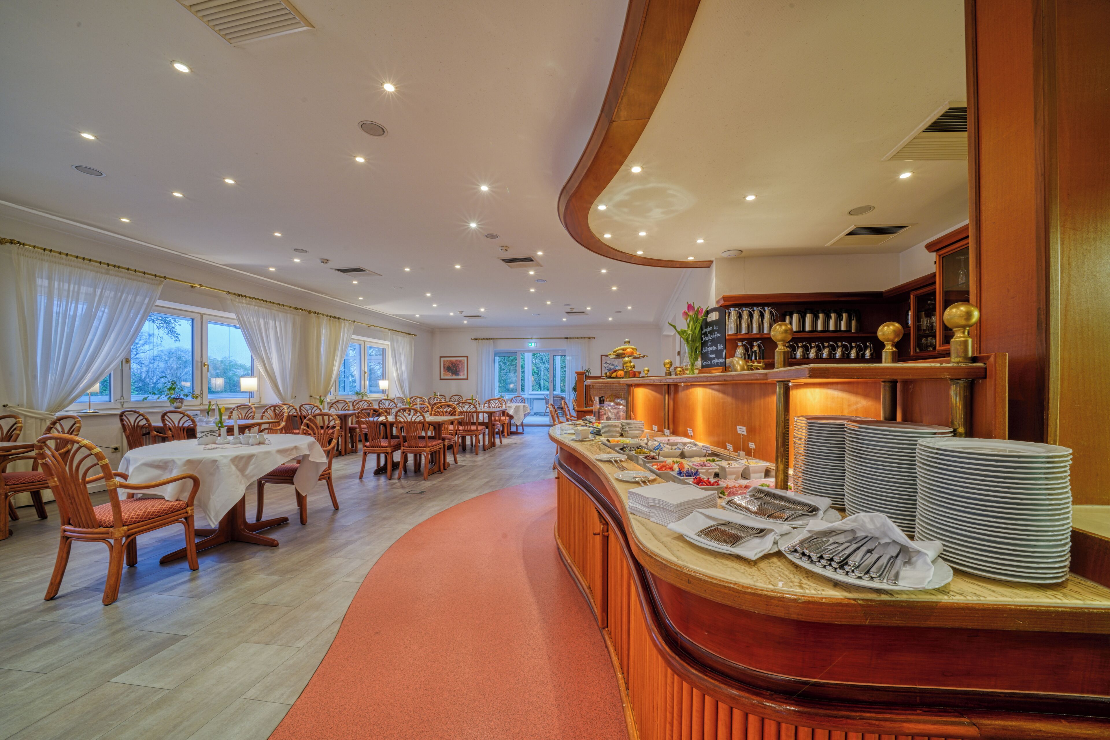 Een restaurantgedeelte van een hotel met ronde tafels en stoelen, een buffet met borden en bestek, en uitzicht op de omgeving door grote ramen.