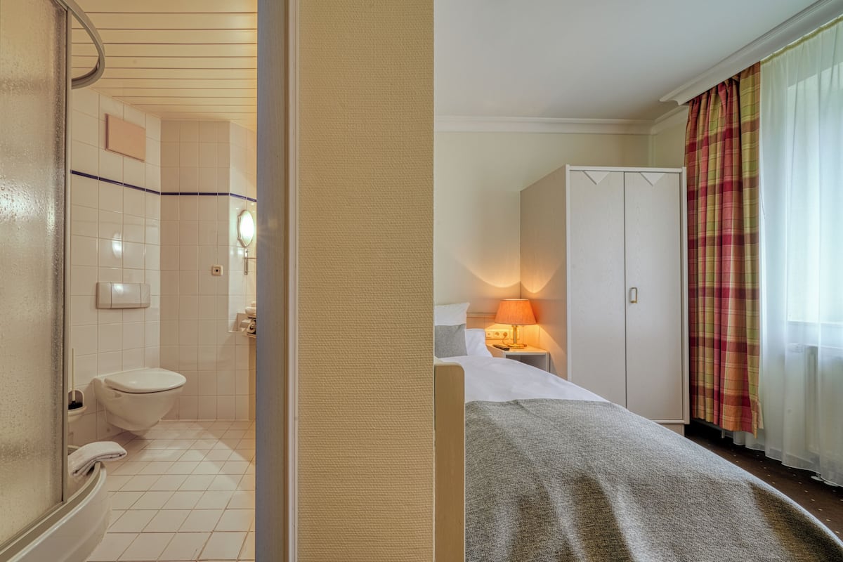 Une salle de bain avec des toilettes, un lavabo et un miroir. La pièce comprend un lit avec une couverture et une table de chevet avec une lampe. Il y a aussi une armoire et une fenêtre avec des rideaux.