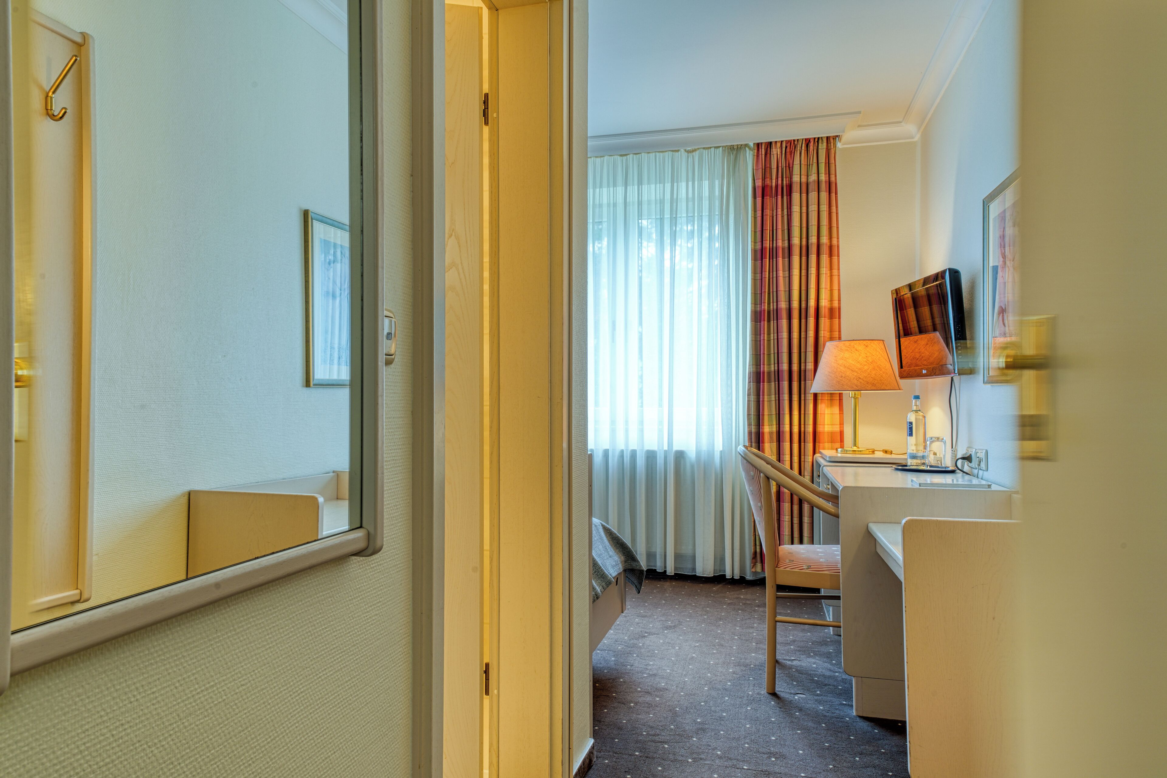A hotel room with a bed, a desk, a chair, a mirror, a window with curtains, and a door.
