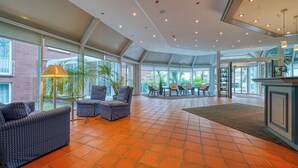 Een ruime binnenruimte met een hoge plafond, grote ramen en diverse zithoekopstellingen, waaronder banken en stoelen.