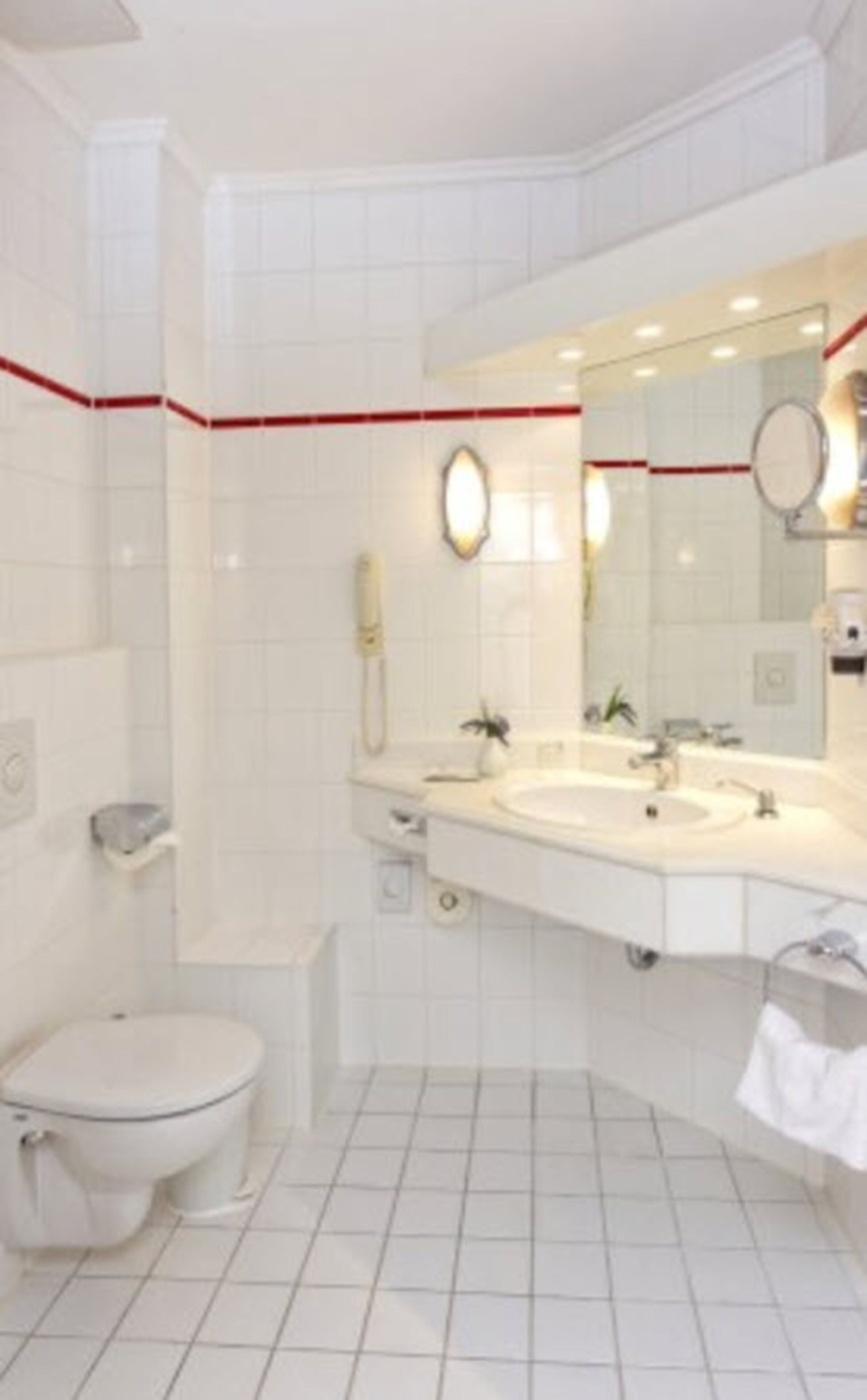 A modern bathroom with a white tiled floor, a large mirror, a double sink, a toilet, and a small shelf with a phone and a plant.