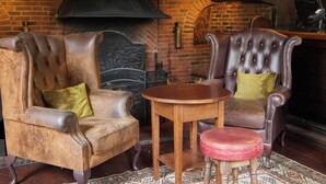 Een knus ingerichte kamer met twee leren fauteuils, een klein houten tafeltje en een rode poef. Een met een bordje bovenop geziene bakstenen openhaard is zichtbaar.