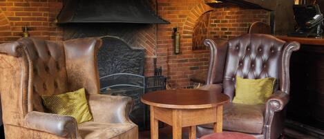 Una habitación acogedora con dos sillones de cuero, una mesita de madera y un reposapiés rojo. Se ve una chimenea de ladrillo con un cartel encima.
