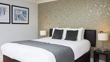 A neatly made bed with white and gray bedding, two framed pictures on the wall, and a patterned wallpaper with birds and leaves.