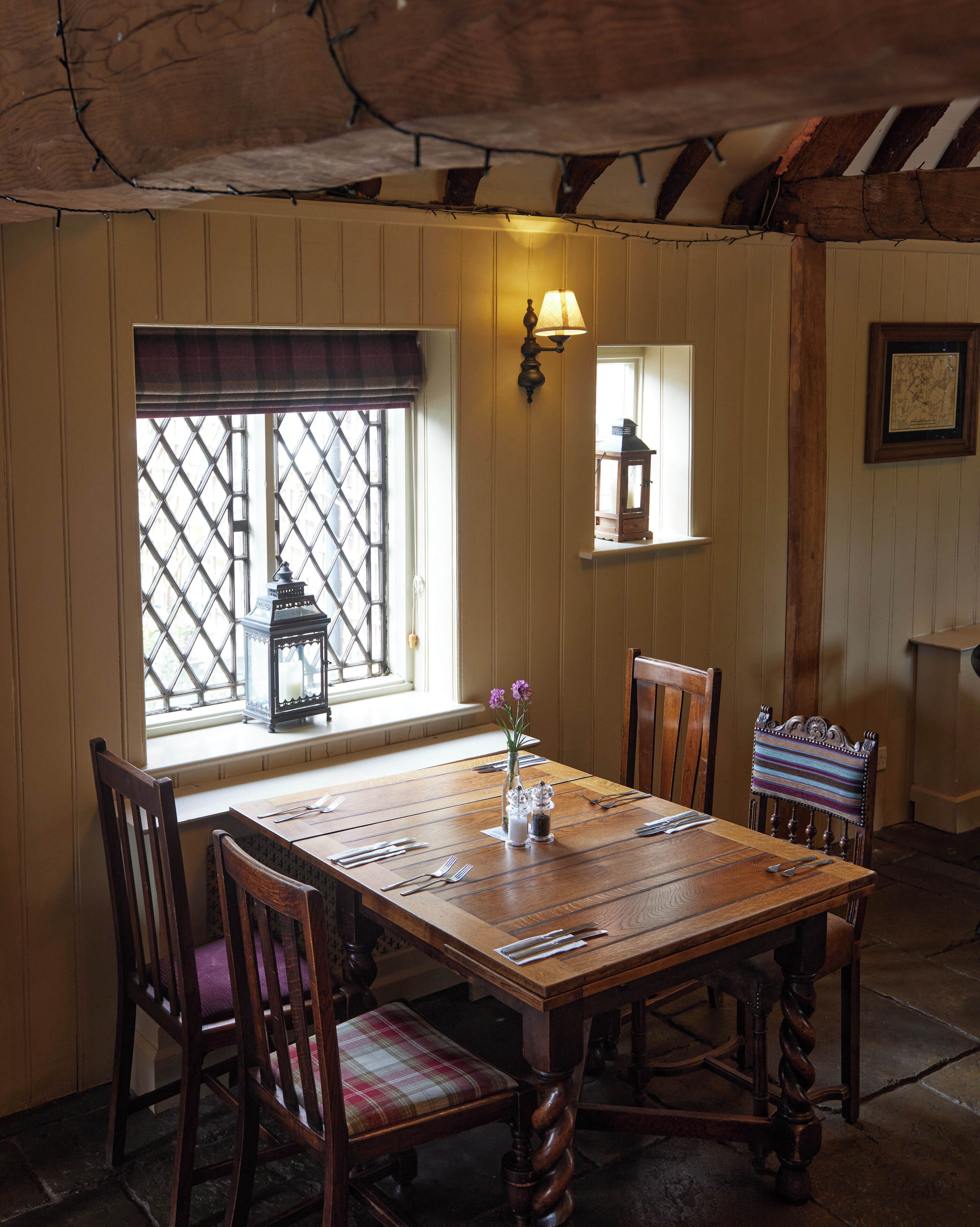 A dining area with a wooden table set for four, surrounded by wooden chairs with cushioned seats. A window with a lattice design allows natural light to enter, and a wall-mounted light fixture provides additional illumination.