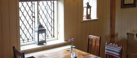 A dining area with a wooden table set for four, surrounded by wooden chairs with cushioned seats. A window with a lattice design allows natural light to enter, and a wall-mounted light fixture provides additional illumination.