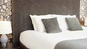 Een netjes opgemaakt bed met witte en grijze kussens, een donker hoofdbord en een lamp op een nachtkastje.