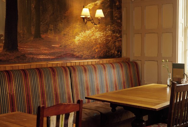 A restaurant with a wooden table and bench seating, a mural of a forest scene, and wall-mounted lamps. - Chequers Inn by Greene King Inns (Uckfield)