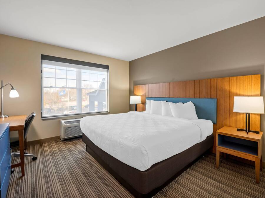 Quarto de hotel com uma cama grande, uma mesa, uma cadeira, duas mesas de cabeceira com abajures, uma janela com vista e um ar condicionado fixado na parede.
