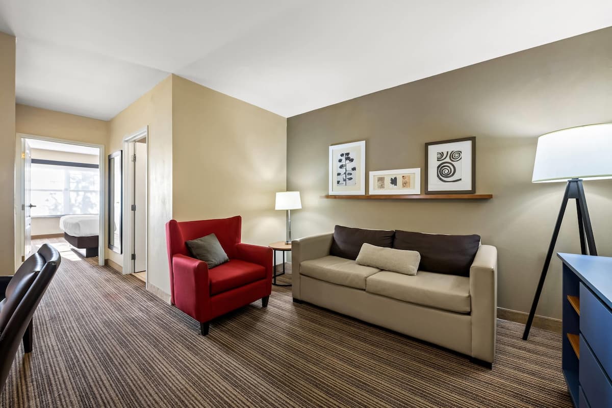 Ein Hotelzimmer mit einem roten Sessel, einem beigen Sofa, einer Lampe und gerahmten Kunstwerken an der Wand.