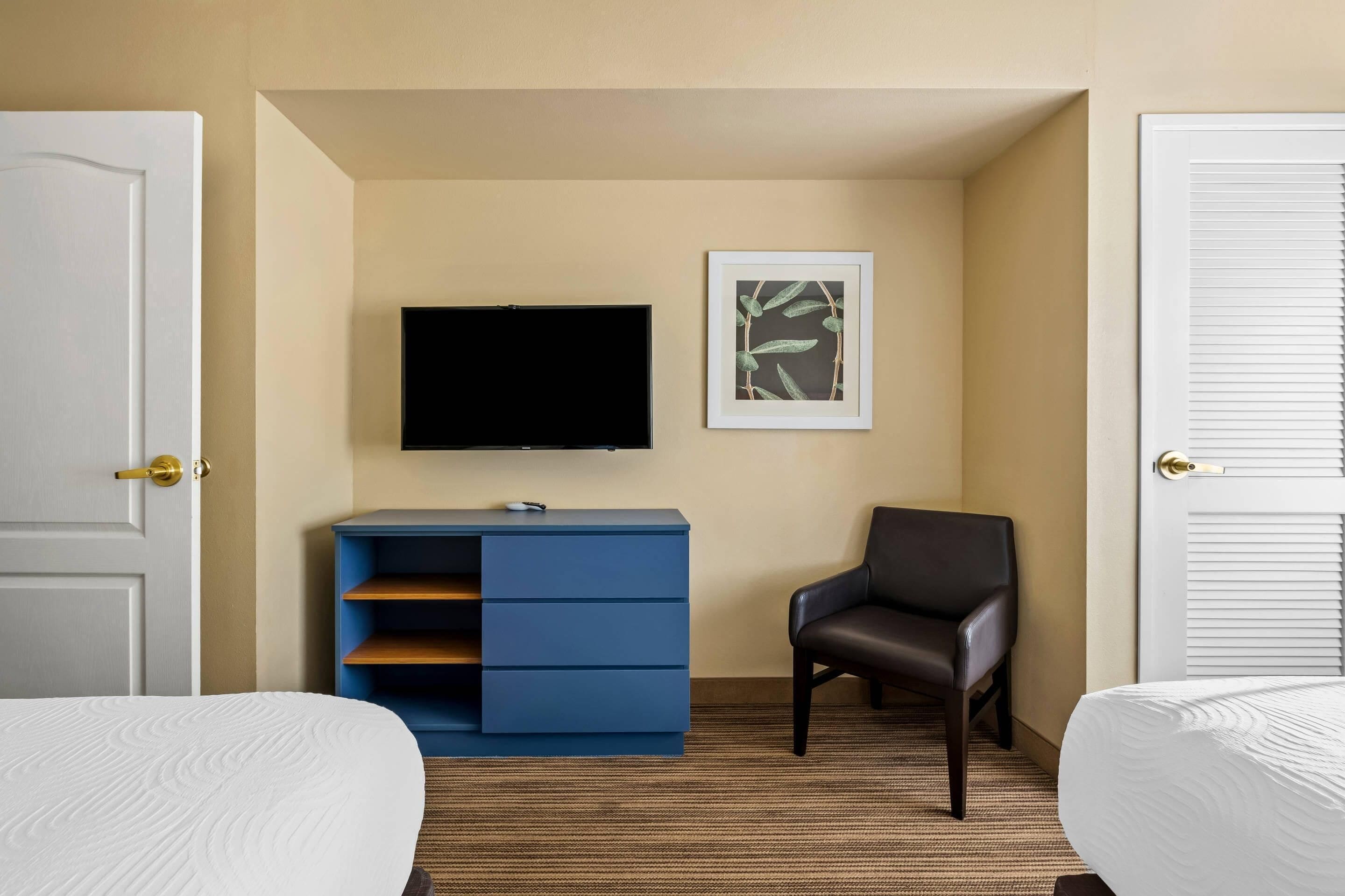Habitació d'hotel amb dos llits, un televisor de pantalla plana, un moble blau, una obra d'art emmarcada i una cadira de pell.