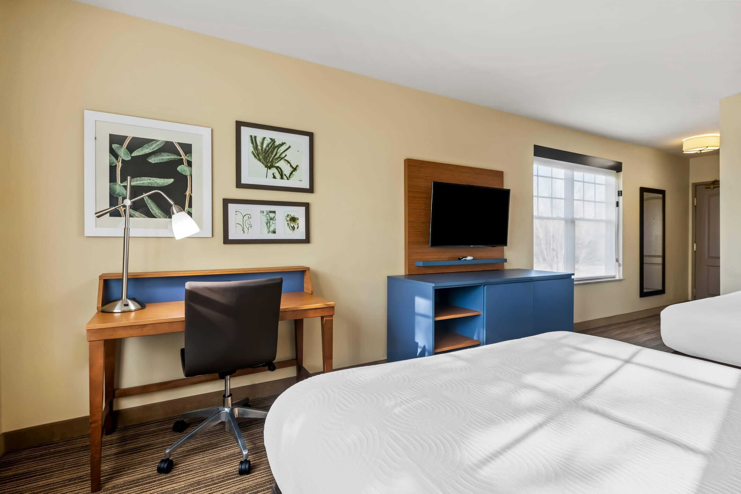 A hotel room with a bed, desk, chair, TV, and artwork on the wall.