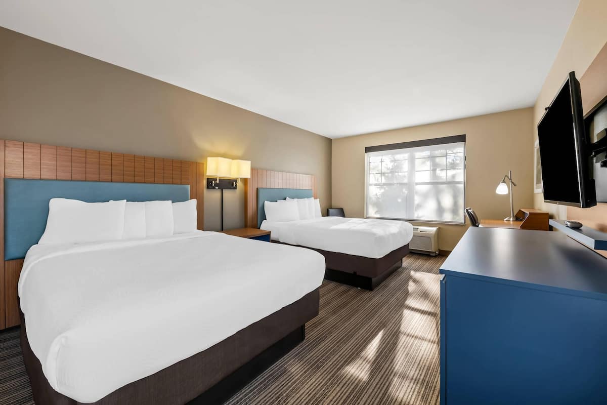 Ein Hotelzimmer mit zwei Betten, einem Schreibtisch, einem Fernseher und einem Fenster.
