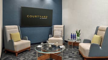 A modern hotel room with two armchairs, a glass coffee table, a small side table with bottles, and a 'COURTYARD BY MARRIOTT' sign on the wall.
