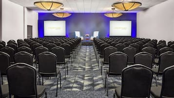 Uma sala de conferências com fileiras de cadeiras, um palco com uma tela e iluminação pendente.