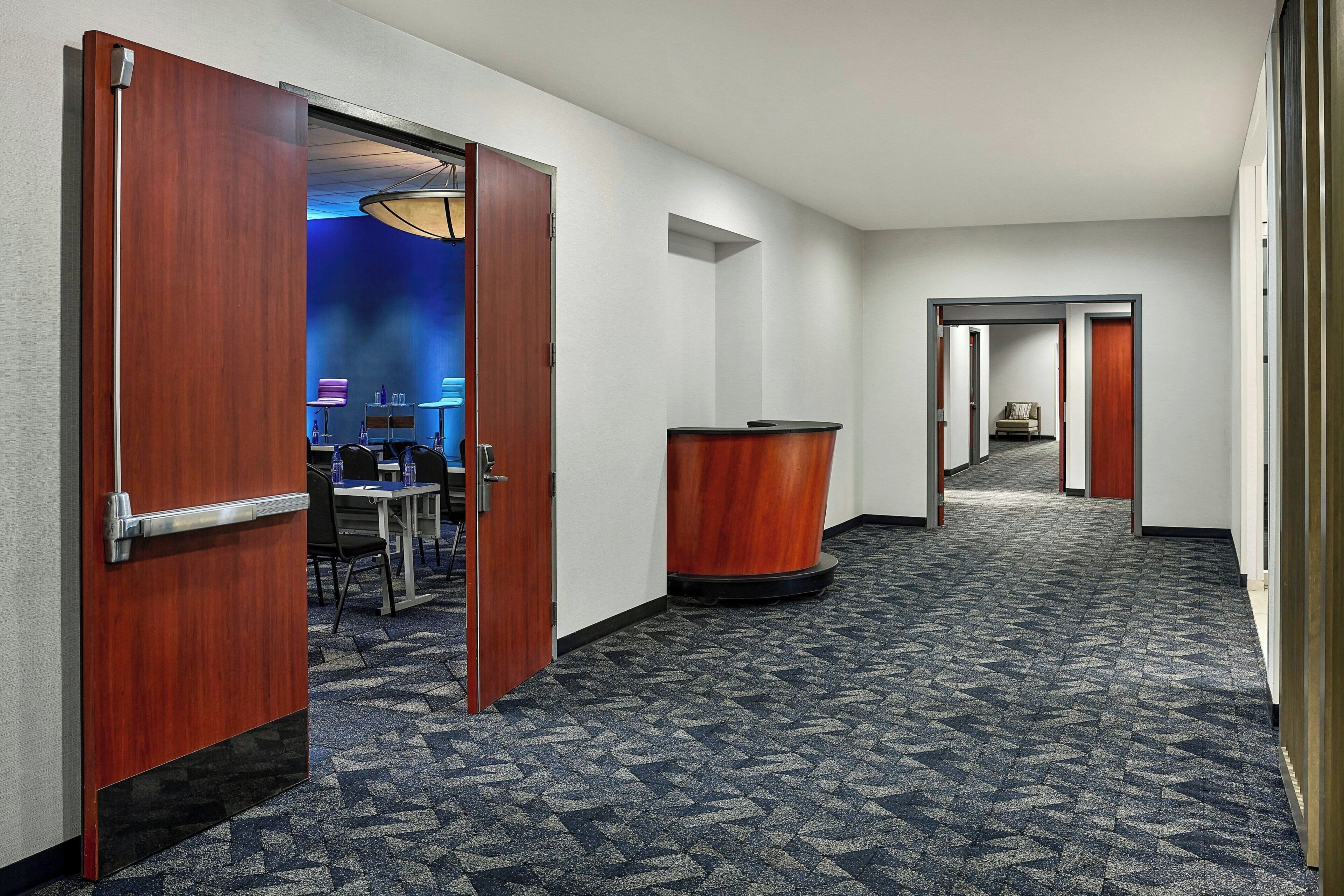 A hotel hallway with a carpeted floor, wooden doors, a reception desk, and a seating area with chairs and tables.