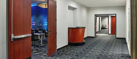 A hotel hallway with a carpeted floor, wooden doors, a reception desk, and a seating area with chairs and tables.