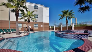 A swimming pool with a tiled edge, palm trees, and a building with a flat roof in the background.