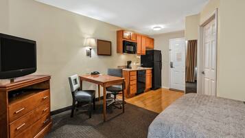 A compact hotel room with a bed, a desk with a chair, a television, a kitchenette with a microwave, refrigerator, and cabinets, and a bathroom visible through an open door.