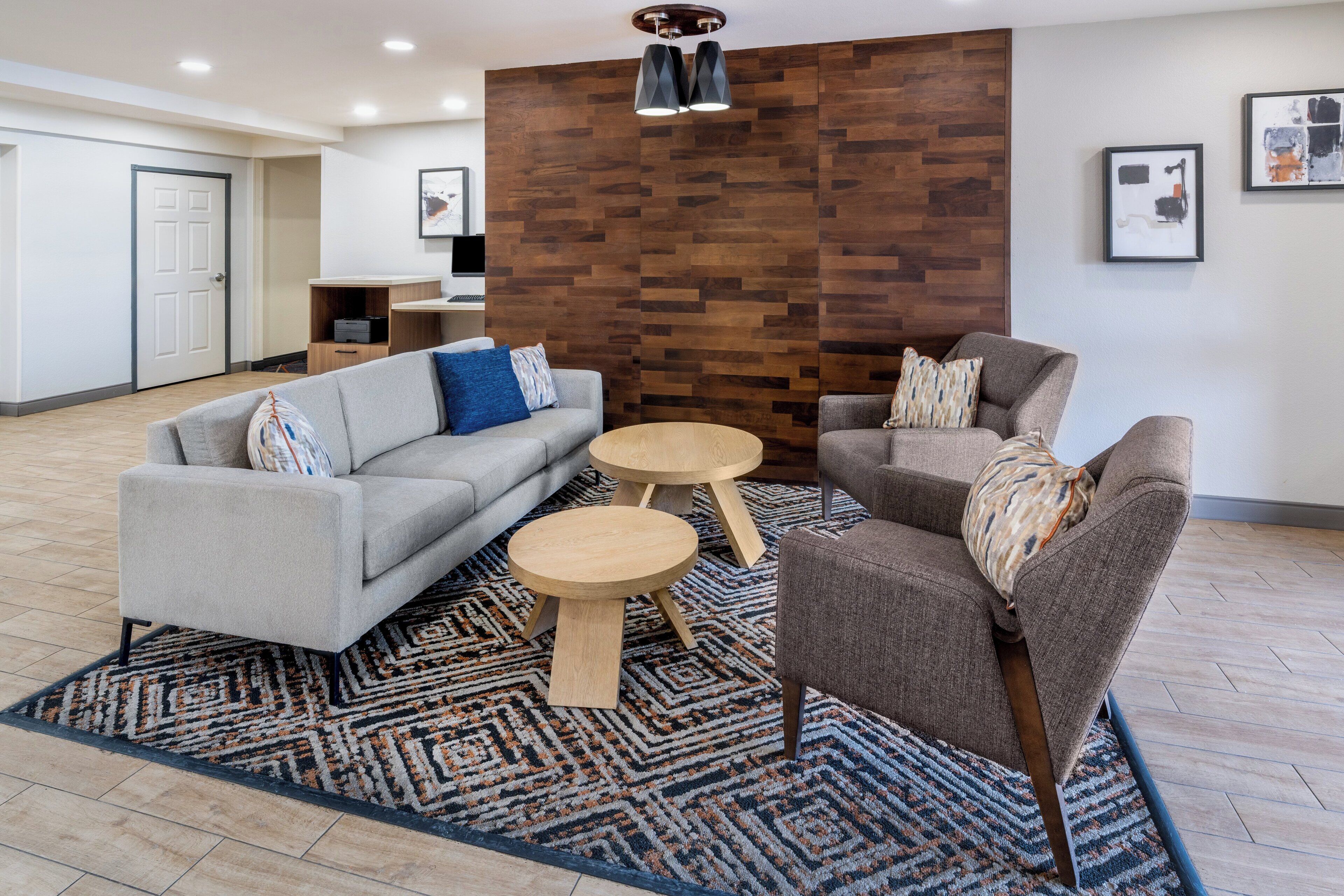 A modern living room with a grey sofa, a brown armchair, and two wooden side tables. A patterned rug covers the floor, and a wooden accent wall is visible behind the furniture.