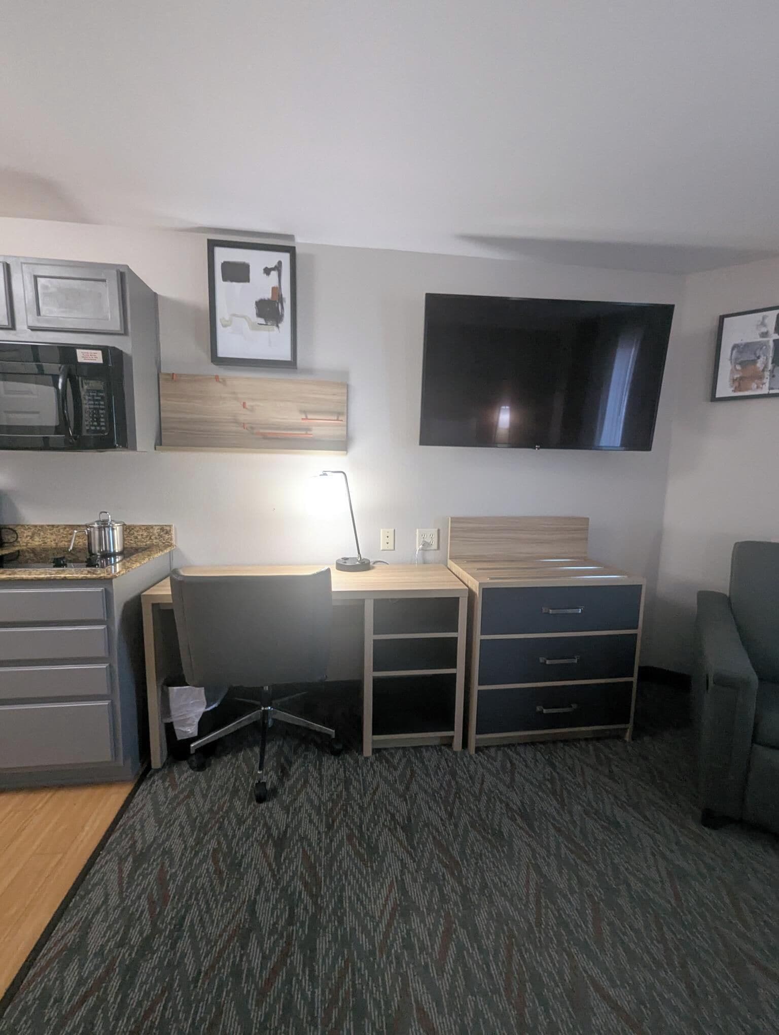 A compact hotel room with a kitchenette, a desk with drawers, a flat-screen TV mounted on the wall, and a gray armchair.