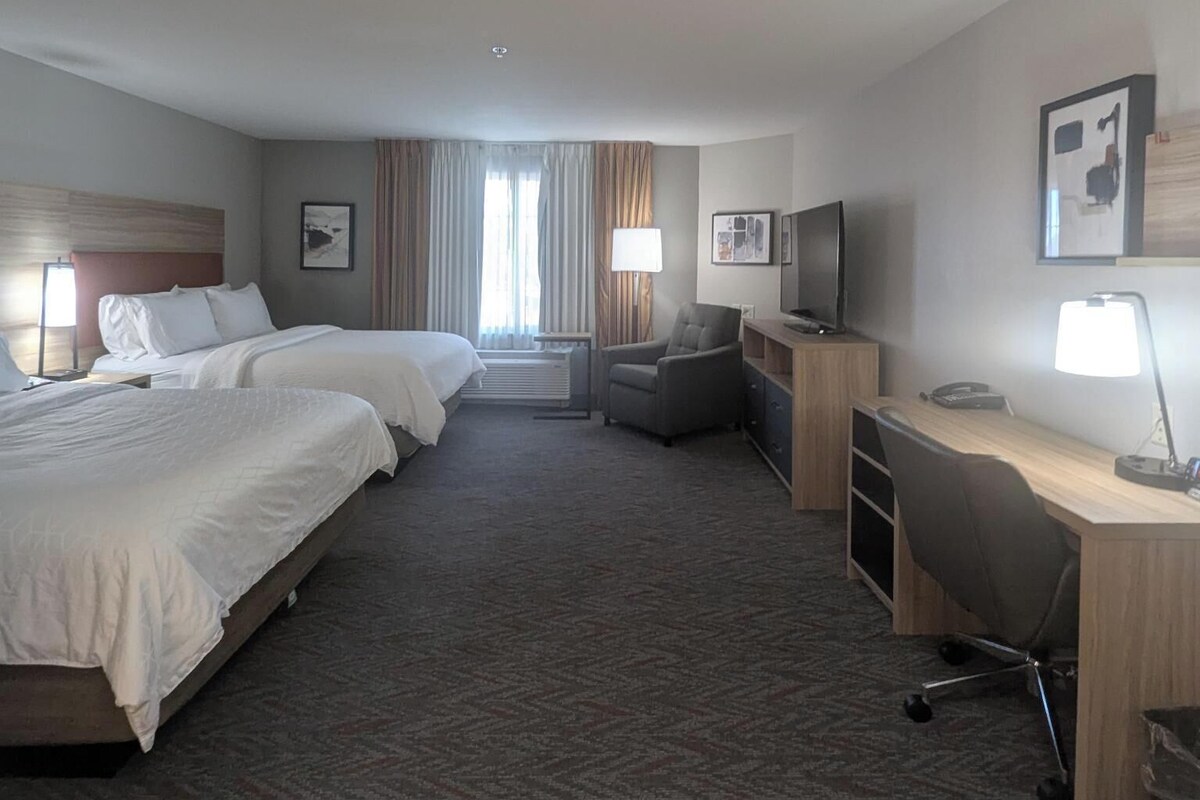 A hotel room with two beds, a desk, a chair, a TV, and a lamp.