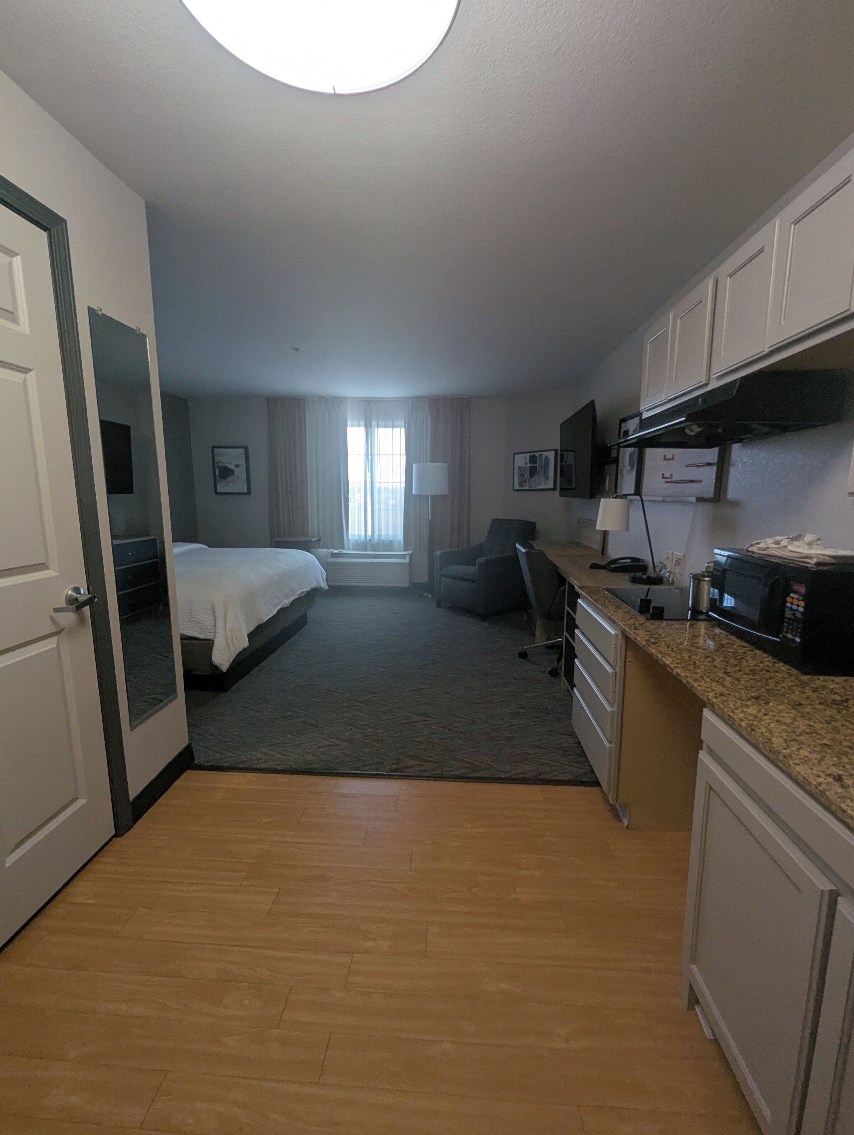 Quarto de hotel com cama, escrivaninha, cadeira, micro-ondas e cozinha compacta.